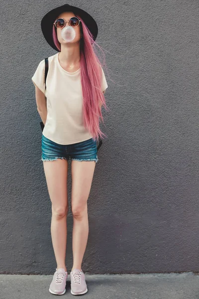 Stylische junge Hipsterfrau mit langen rosafarbenen Haaren, die eine Blase mit Kaugummi pustet. — Stockfoto