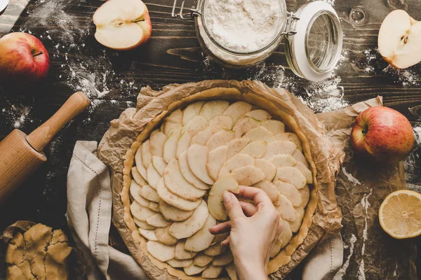 Cooking apple pie. Hands adding apple slices to pie crust. Apple pie ingredients on wooden table.