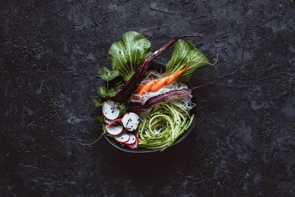 Raw asian bowl with rice noodles, carrot, radish and cucumber on black background. Top view.