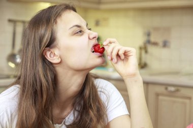 Güzel genç kız meyve ve taze çilek yiyor mutfakta, çilek ısırma