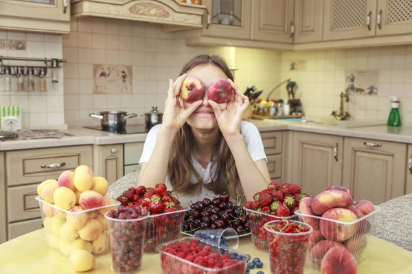 Beautiful young girl eats fruits and fresh berries in the kitchen, eyes are covered with peaches