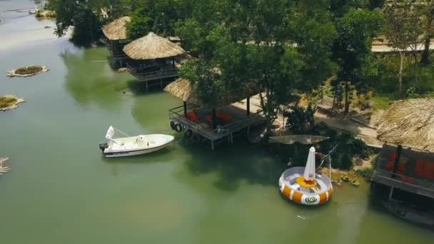 Bungalows d'été avec toit de chaume et bateau dans le lac vert dans l'eau villa station vue sur drone. Les gens nagent dans les eaux vertes lac de montagne vue aérienne . — Video