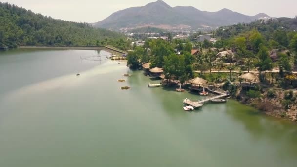 Vista aérea casas de verão com telhado de palha e cais de barco na costa em lago de montanha entre a natureza tropical para descansar. Bela paisagem montanhosa e água verde na vista lago drone . — Vídeo de Stock