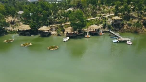 Bungalows en bois avec toit de chaume et jetée de bateau sur le rivage lac vert dans hôtel station vue sur drone. Chalets au bord du lac de montagne au milieu de la nature sauvage dans l'eau villa station paysage aérien . — Video