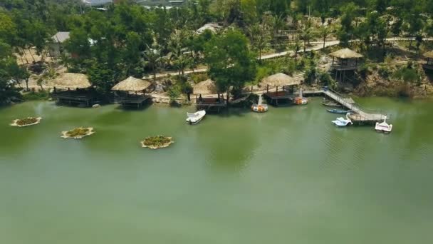 Holzbungalows mit Reetdach und Bootsanlegestelle am Ufer des grünen Sees im Drohnenblick des Resorthotels. Ferienhäuser am Ufer Bergsee inmitten wilder Natur in Wasser Villa Resort Antennenlandschaft. — Stockvideo