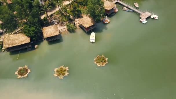 Summer bungalows with thatched roof and boat pier on shore green lake in luxury mountain hotel aerial view. Cottages on shore tropical lake among exotic nature in water villa resort drone view. — Stock Video