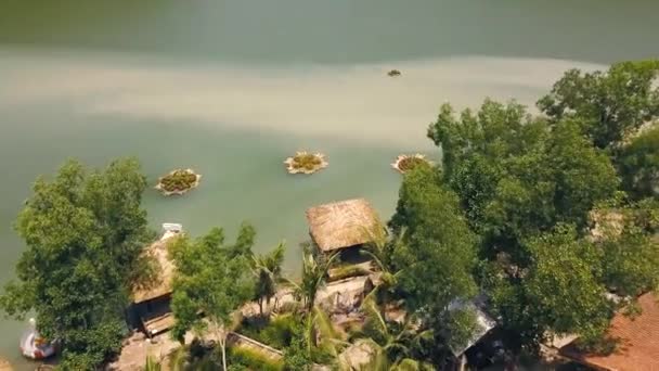 Tropical bungalows with thatched roof and boat pier on shore green lake in resort hotel mountain aerial view. Cottages on shore mountain lake among exotic nature in water villa resort drone view. — Stock Video