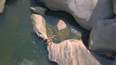 Nehir büyük taş ve su hava görünümünde daldırma ayak üzerinde oturan bir genç bayan. Mutlu kadın ayaklarını clea nehir su dron görünümünde ıslak.