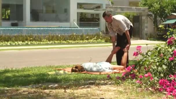Ταϊλανδικό massagist κάνει μασάζ σε γυναίκα ξαπλωμένη για την εξωτερική πράσινη χλόη. Επαγγελματική massagist ανθρώπου κάνει παραδοσιακή γιόγκα μασάζ. Έννοια της εναλλακτικής ιατρικής. — Αρχείο Βίντεο