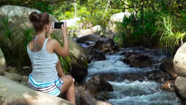 Hermosa mujer haciendo fotos en el teléfono móvil mientras viaja en cascada de montaña en el bosque selvático. Mujer turista disparo de vídeo en el teléfono inteligente mientras sube om río de montaña — Vídeo de stock