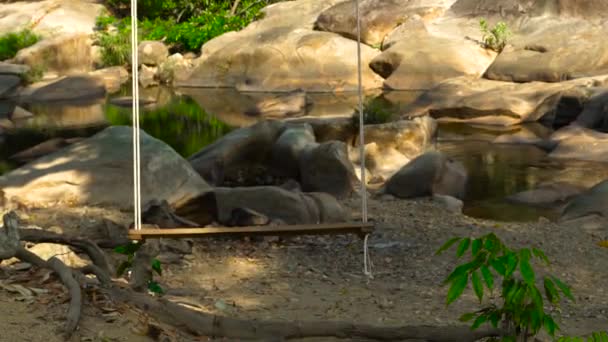 Balancez sur la rivière de fond et le rivage pierreux. Balançoire en bois balançant sur le rivage de la rivière sur fond beau paysage rochers dans l'eau . — Video