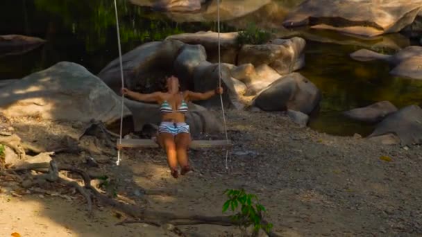 海岸の石の多い川でスイングに若い女性は。背景のジャングルの風景川岸の揺れを楽しんで幸せな女。自由と空想のコンセプト. — ストック動画