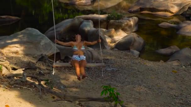 Mujer joven en columpio en la orilla del río pedregoso. Mujer feliz disfrutando balanceándose en la orilla del río en el paisaje de la selva de fondo. Libertad y concepto de ensueño . — Vídeos de Stock