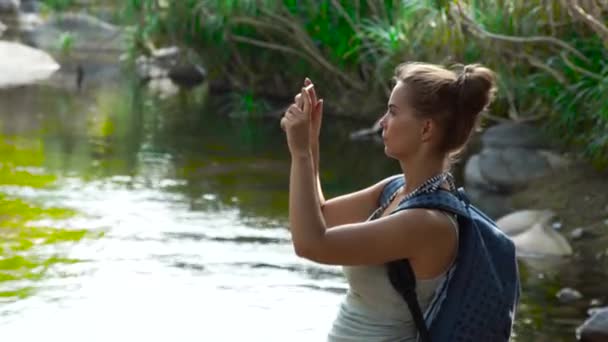 Traveling woman using mobile phone for photo while hiking in jungle forest. Woman tourist shooting video on smartphone while climbing on river in rainforest. — Stock Video