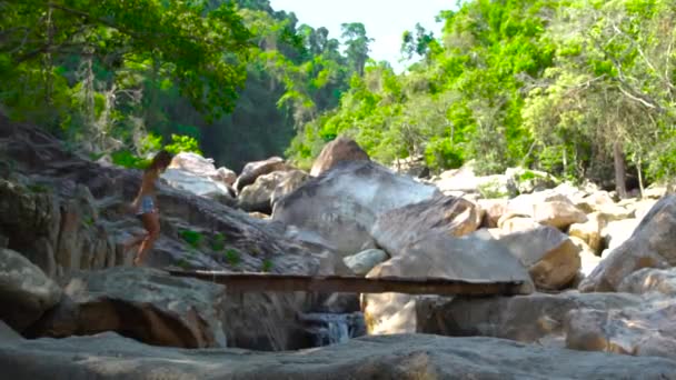 Schöne Frau, die auf einer hölzernen Brücke über den schnellen Fluss in den Bergen geht. Touristin wandert auf Brücke über Berge und steinigen Fluss im Dschungel. — Stockvideo