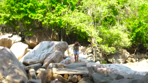 Mujer turística caminando en el puente sobre el río de montaña durante el senderismo de verano. Mujer joven caminando sobre el puente del río en el bosque tropical. Concepto de actividades de verano . — Vídeos de Stock