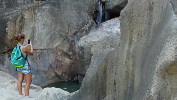 Mujer viajera haciendo fotos en un teléfono inteligente mientras escalaba en una cascada de montaña en la selva tropical. Mujer turista filmando vídeo en el teléfono móvil mientras camina en el río de montaña . — Vídeos de Stock