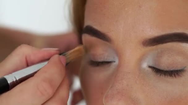 关闭 visagiste 使用化妆刷的应用 eyeshadows 在眼睑化妆模型。化妆艺术家应用魅力化妆的女人的脸。美容时尚产业. — 图库视频影像