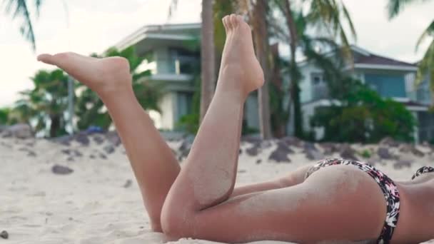 Gebruinde vrouw liggend op zandstrand en benen zwaaien. Billen en benen sexy vrouw onder zand dicht omhoog. Aantrekkelijke vrouw zonnebaden op het strand van de zomer. — Stockvideo