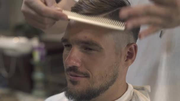 Ansikte stilig man samtidigt kamma hår och skägg i barbershop. Närbild av haircutter kamma håret att skäggig man i manliga salong. Hipster frisyr koncept. — Stockvideo