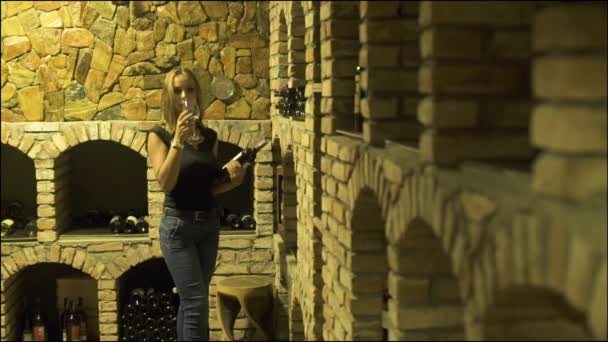 Женщина пьет красное вино из бокала и смотрит на бутылку в подвале. Женщина винодел дегустации и пить красное вино из бокала в традиционном подвале . — стоковое видео