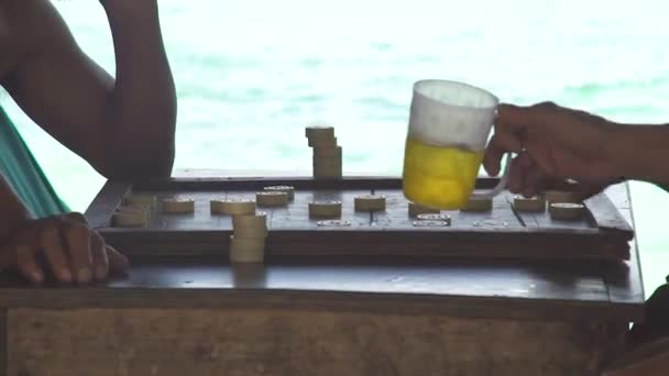 Män spelar backgammon och dricka öl från mugg på havsstranden i skugga. Män som spelar backgammon styrelsen med öl glas på blå havet landskapet. — Stockvideo