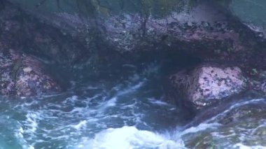 Kayalık adada sıçramasına dalgalar mavi deniz. Dalgalar üzerinde kayalık uçurum kırma köpük. Şeffaf deniz suyu ve büyük taşlar arka plan.