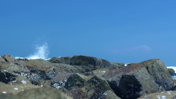 Stones on sea shore and splashing water waves on blue sky landscape. Water waves breaking at rocky beach, ocean shore on blue sky background. — Stock Video