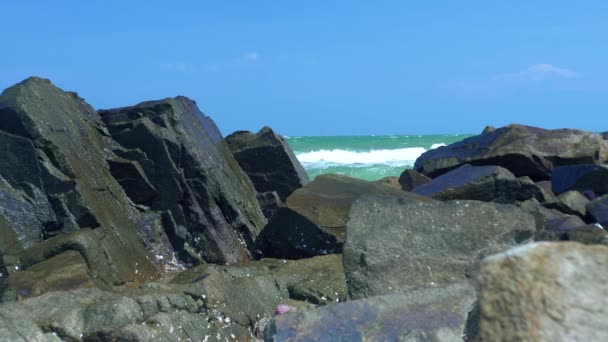 Rocky shore and splashing sea water on blue sky landscape. Sea waves crushing at stony beach. Water waves breaking on stony coast in ocean on blue sky background. — Stock Video