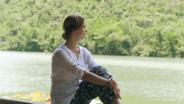 Chica disfrutando de verano río y montaña cubierto de bosque verde. Mujer joven relajándose en la orilla del lago y mirando verde paisaje de montaña . — Vídeo de stock