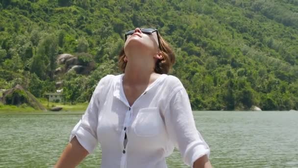 Ευτυχισμένη γυναίκα απολαμβάνοντας ποταμό και το βουνό καλύπτεται καταπράσινο δάσος. Όμορφο κορίτσι χαλάρωση στην όχθη της λίμνης και να ψάχνει στον ουρανό σε πράσινο ορεινό τοπίο. — Αρχείο Βίντεο