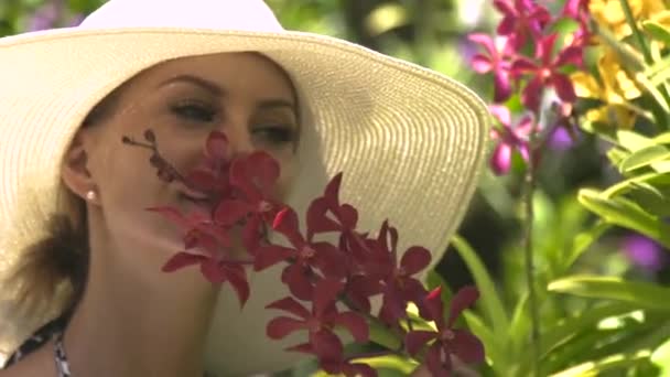 Mujer joven en sombrero disfrutando de flores en flor en el jardín de verano. Chica oliendo aroma de orquídeas florecientes en el jardín de flores en el día soleado . — Vídeo de stock