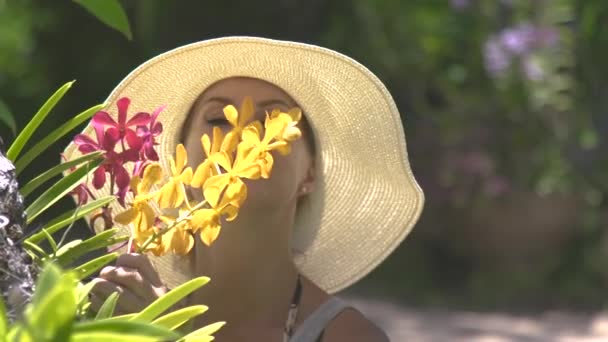 Beautiful woman sniffing aroma blooming flowers in summer garden. Girl in hat enjoying scent of blooming orchids in garden at sunny day.