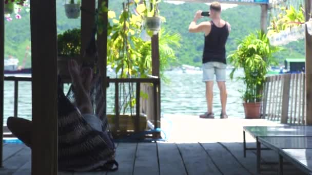 Ludzie w hamak lato na brzegu morza i fotografowanie krajobrazu do telefonu komórkowego. Człowiek, co morze zdjęcie do smartfona na odkryty taras z hamakiem. — Wideo stockowe