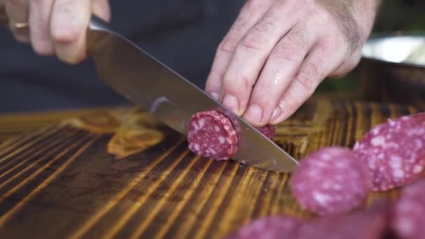 木の板にナイフ切断ソーセージをクローズ アップ 台所のテーブルのサラミ ソーセージをスライス ナイフで男性の手 イタリアのピザのための成分 肉珍味とスナック 料理プロセス — ストック動画