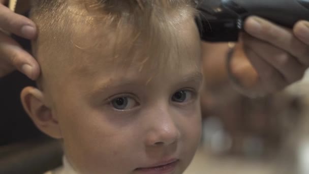 Retrato menino enquanto cortando com navalha elétrica na barbearia de perto. Crianças conceito de corte de cabelo. Crianças cabeleireiro cortar cabelo para menino com barbeador elétrico no salão de barbeiro . — Vídeo de Stock