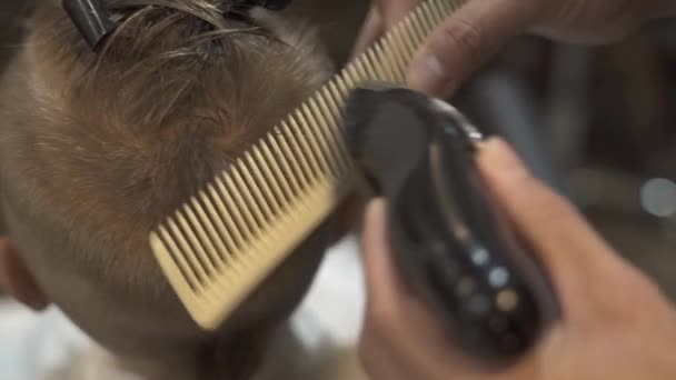 Cortar el cabello de los niños con afeitadora eléctrica y peinar en la barbería. Primer corte de pelo de niño en el salón de niños. Chico corte de pelo con peine y afeitadora eléctrica. Peluquero haciendo peinado chico . — Vídeos de Stock