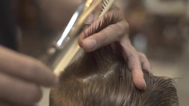 Hairdresser combing and cutting wet hair with barber scissors in male salon close up. Children haircut with hair scissors. Little boy hairdressing in barber shop. Kid haircut concept.
