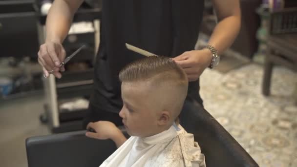 Hairdresser cutting wet hair of little boy with barber scissors in male salon. Children haircut with scissors in hairdressing salon. Little boy hairstyle in barber shop. Kid cutting hair.