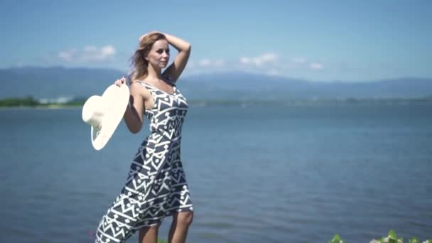 Hermosa mujer joven en vestido y sombrero de pie en el mar y las montañas paisaje. Mujer atractiva con sombrero posando en la orilla del río, montañas y fondo azul del cielo . — Vídeo de stock