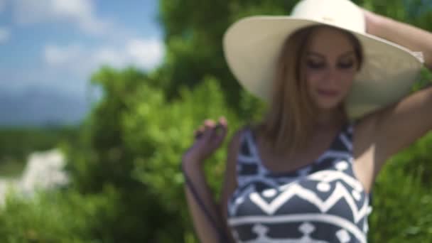 Mooie jonge vrouw in jurk en muts op groene bomen achtergrond op zomerdag. De aantrekkelijke vrouw portret in de hoed op zomer bomen achtergrond. — Stockvideo