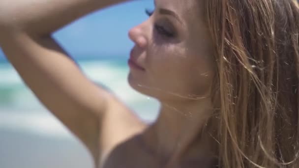Mooie jonge vrouw aanraken van de lucht en op zoek naar camera op zomer strand op blauwe zee achtergrond. Close-up van portret gelukkig aantrekkelijke vrouw in bikini op strand. — Stockvideo