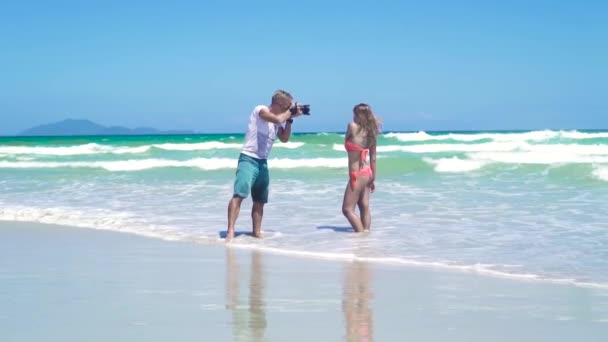 Homem fotógrafo tirar fotos de jovem mulher de biquíni posando na praia do mar. Jovem fotógrafo trabalhando com mulher modelo no mar. Homem usando câmera de fotos para sessão de fotos de mulher bonita . — Vídeo de Stock