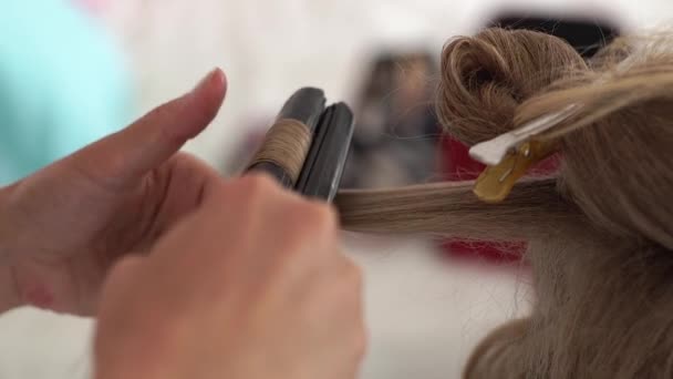 Peluquero usando pinzas para rizar el cabello de cerca. Proceso de creación de peinado y rizado pelo largo con plancha para el cabello en el estudio de belleza. De cerca. Peluquería haciendo peinado con estilo a la mujer rubia — Vídeos de Stock