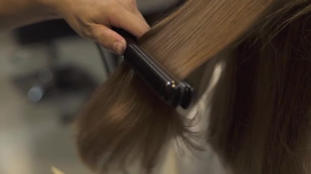 Stylist straightening long hair with iron and combing while hairstyling in hairdressing salon. Woman hairstyling with hair iron in beauty studio. Master creating female hairstyle in barbershop. — Stock Video