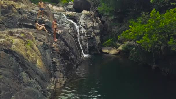 Man jumping in mountain river from waterfall in tropical forest drone view. People diving in river water flowing from mountain waterfall in rainforest. — Stock Video