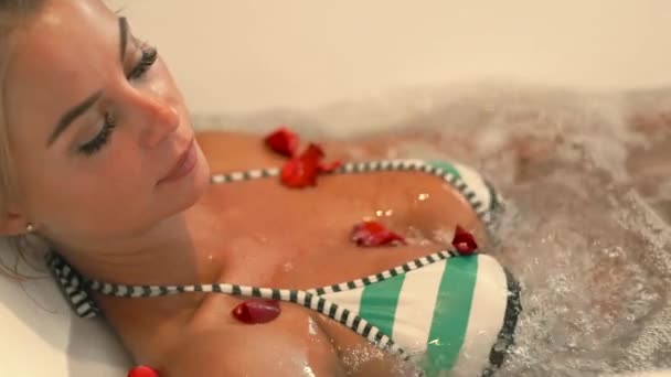 Bella donna in bikini che fa il bagno jacuzzi con petali di rosa nella spa. Viso ragazza sdraiata nella vasca idromassaggio nel resort di lusso spa. Terapia estetica e concetto di cura del corpo. Concetto bagno romantico — Video Stock