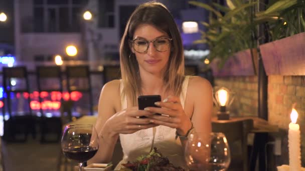 优雅的女人用手机坐在晚上餐厅的餐桌上。美丽的女人看智能手机在晚上晚餐在餐厅. — 图库视频影像