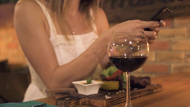 Mujer joven usando smartphone mientras cena en el restaurante de barbacoa. Mujer navegando redes por teléfono móvil en la cena de la noche en restaurante de carne . — Vídeos de Stock