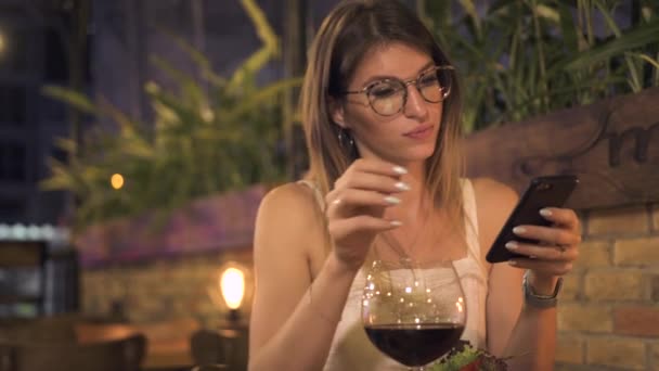 Mooie vrouw chatten in sociale netwerken en het drinken van rode wijn in de avond restaurant. Jonge vrouw met behulp van smartphone en drinken uit wijnglas bij het diner in restaurant. — Stockvideo